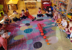 Dzieci siedzą na dywanie. Po środku ustawione dynie od największej do najmniejszej.